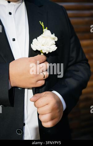 Gesichtsloser Bräutigam in schwarzem Smoking und weißem Hemd hält eine weiße Nelken-Boutonniere nahe an seiner Brust, sein goldener Ehering ist an der Hand sichtbar. Stockfoto