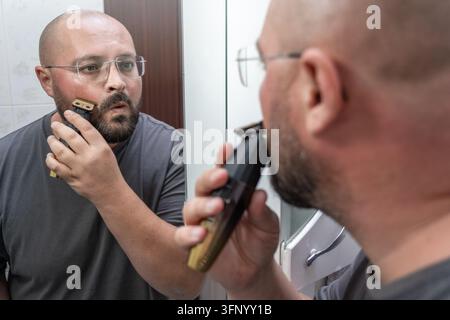 Mann trimmt Bart mit elektrischem Trimmer, während er in den Badezimmerspiegel schaut, die Wangenlinie und Gesichtsbehaarung für präzise Pflege, Selbstpflege und eine ordentliche Home-Barber-Routine formt. Stockfoto
