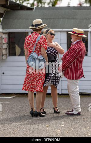Drei Goodwood-Revival-Teilnehmer in historischer Kleidung, die neben Holzhütten am Strand plaudern, mit geblümten Kleid, gepunktetem Kleid und gestreiftem Blazer. Stockfoto