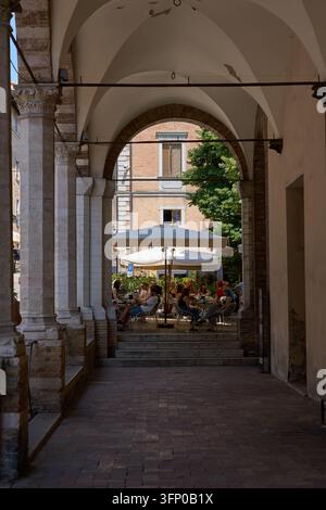 Urbino, Italien - 21. Juni 2026 - Blick durch einen historischen Steinbogen zu einer Außenterrasse eines Cafés auf einem sonnigen italienischen Stadtplatz Stockfoto