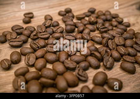 Verstreute geröstete Kaffeebohnen bedecken eine warme Holzoberfläche, zeigen verschiedene Brauntöne, gespaltene Nähte, abgerundete Formen und natürliche Textur Stockfoto