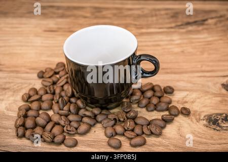 Leere glänzende schwarze Kaffeetasse, umgeben von gerösteten Kaffeebohnen auf einer warmen Holzplatte in einem einfachen rustikalen Stillleben-Arrangement Stockfoto