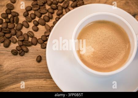 Weiße Keramiktasse mit Espresso und goldener Crema sowie verstreuten gerösteten Kaffeebohnen auf einem rustikalen Holztisch in warmem natürlichem Licht Stockfoto