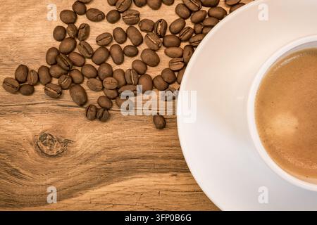 Oberblick auf eine weiße Keramiktasse gefüllt mit Kaffee und Crema neben verstreuten gerösteten Bohnen auf einem rustikalen Holztisch, mit warmem Braunton Stockfoto