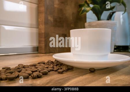 Weiße Kaffeetasse und Untertasse, umgeben von gerösteten Kaffeebohnen auf einem Holztisch, mit einer Topfpflanze und warmem Zuhause-Interieur Stockfoto