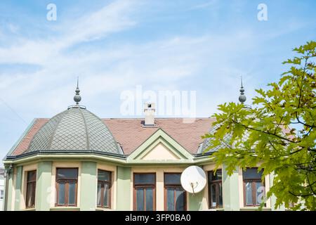 Verzierte Gebäudefassade mit zwei kuppelförmigen Dachtürmen, gemusterten Ziegeln, Schornstein, Satellitenschüssel, laubigem Baum und blauem Himmel bei hellem Tageslicht Stockfoto