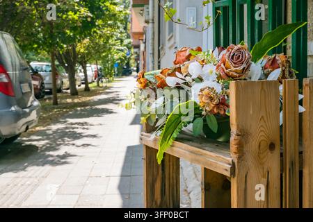 Buntes Arrangement aus künstlichen Blumen, das in einem rustikalen hölzernen Straßenstand neben einem sonnigen, von Bäumen gesäumten Wohnweg und einem geparkten Auto ausgestellt ist Stockfoto