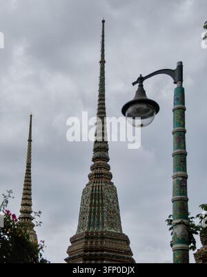 Perspektivaufnahme mehrerer Tempelturmspitzen, die hinter einem historischen geschwungenen Laternenpfahl im Innenhof des Wat Pho-Tempelkomplexes in Bangkok, Thailand, ausgerichtet sind. Stockfoto