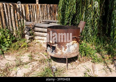 Ein alter, verwitterter und rostiger handgefertigter Metallkesselofen steht auf dem Boden in einem ländlichen Hinterhof Stockfoto