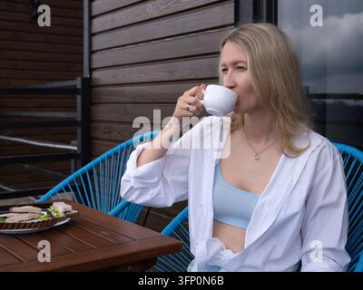 Frau trinkt gemütlich Kaffee aus einer weißen Tasse, während sie auf einem Balkon sitzt, und genießt ein ruhiges Frühstück im Freien mit offenen Sandwiches, um Entspannung zu genießen Stockfoto