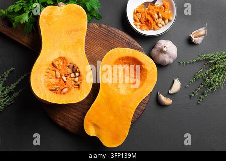 Rohe Kürbis Hälften oder Butternusskürbis auf Schneidebrett, Kräuter, Knoblauch auf schwarzem Hintergrund, Vogelperspektive. Herbstliches Kochkonzept. Stockfoto