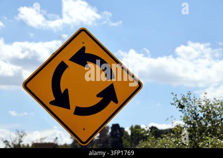 Ein rautenförmiges gelbes Warnschild mit schwarzen kreisförmigen Pfeilen, das auf einen kreisrunden Verkehrskreisel voraus hinweist. Stockfoto
