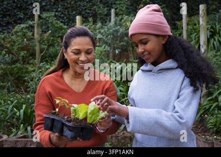 Afroamerikanische Mutter hält ein schwarzes Saadtray, während ein Kind mit rosa Mütze Pflanzen im Hinterhof überprüft Stockfoto