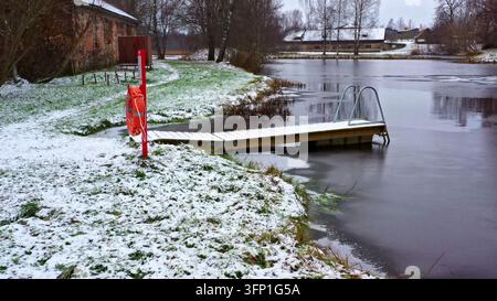 Luftaufnahme eines hölzernen Stegs, der sich in einen teilweise zugefrorenen Teich erstreckt, umgeben von schneebedecktem Gras und einem historischen Backsteingebäude in Lettland. Stockfoto
