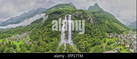 Luftbild der Acquafraggia-Wasserfälle, die einen üppigen grünen Berghang hinunterstürzen, umgeben von einem kleinen Dorf und nebligen Gipfeln in der Lombardei, Italien. Stockfoto