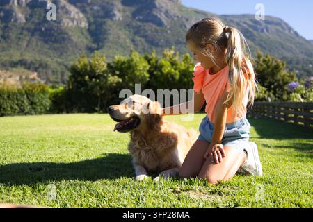 Mädchen in pinkfarbenem T-Shirt und Jeansshorts sitzt auf dem Rasen und streichelt einen Golden Retriever, Kopierraum Stockfoto