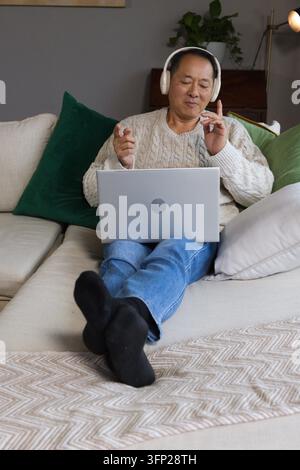 Asiatischer Senior entspannt sich auf einem Ecksofa zu Hause, trägt einen cremefarbenen Pullover und nutzt einen Laptop mit Kopfhörern Stockfoto