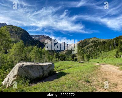 Blick auf den Uinta National Forest mit einem großen Felsbrocken, einem Schotterweg, einer grünen Wiese und Espen Bäumen vor Bergen unter einem blauen Himmel in Utah County, Ut Stockfoto