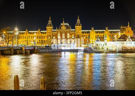 Blick auf die beleuchtete Fassade des Amsterdam Centraal Bahnhofs, die sich nachts auf dem Wasser in Amsterdam, Nordholland, Niederlande, spiegelt. Stockfoto