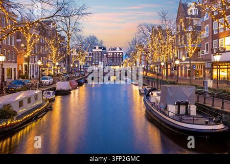 Blick auf einen Kanal in der Dämmerung mit festlichen Lichtern an den Bäumen, historischen Gebäuden und Booten, die entlang der Ufer in Amsterdam, Nordholland, Niederlande, festgemacht sind. Stockfoto