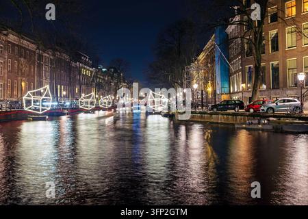Ansicht von beleuchteten Lichtkunstinstallationen, die nachts auf einem Kanal schweben, mit historischen Kanäushäusern und Reflexionen im Wasser in Amsterdam, Norden Stockfoto