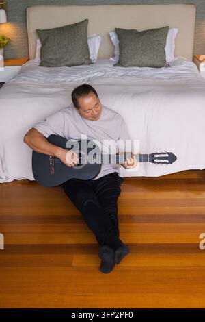 Seniorer asiatischer Mann sitzt im Schlafzimmer auf poliertem Holzboden und spielt schwarze klassische Gitarre Stockfoto