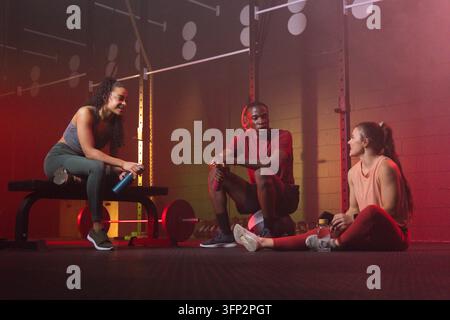 Verschiedene Fitnessstudio-Freunde, die zwischen den Sätzen im Gewichtsaal mit Hantelstange, Wasserflaschen, Kopfhörern ruhen. Fitness, Training, Trio, lässig, Ausrüstung, Bank Stockfoto