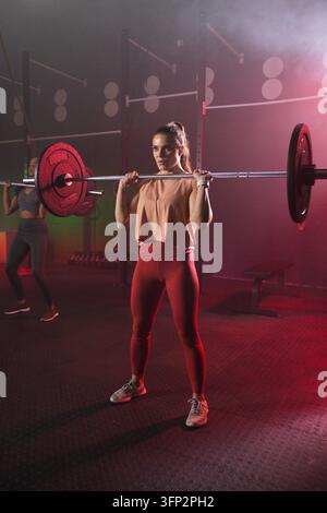 Zwei Frauen heben mit roten Gewichten beladene Hantelstangen im Fitnessstudio, tragen ein pfirsichfarbenes Oberteil und rote Leggings Stockfoto