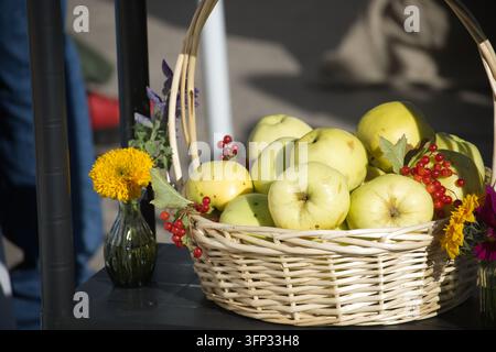 Ein rustikaler gewebter Behälter hält geerntete Obstgartenfrüchte, akzentuiert durch lebendige Wildblumen in kleinen Glasvasen während eines hellen Outdoor-Ernteereignisses Stockfoto