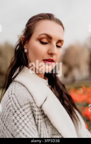 Frauenporträt Eleganz. Gelassene Frau in einem grauen Wollmantel mit Pelzkragen steht in einem Herbstpark mit orangefarbenen Blumen Stockfoto