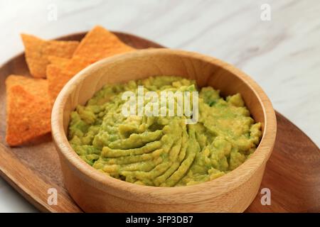 Frische Guacamole in einer Holzschüssel mit Tortilla-Chips. Die hellgrüne Textur lädt zu gesundem Naschen ein. Das Holztablett verleiht eine rustikale, warme Anziehungskraft. Perfekt für h Stockfoto