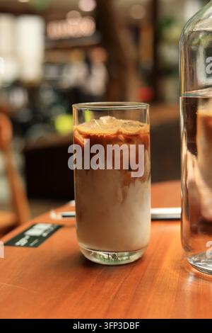 Ein Glas Eiskaffee steht auf einem warmen Holztisch. Die Zuckerschicht wirbelt sanft, während das Getränk langsam abkühlt. Der weiche Hintergrund deutet auf eine gemütliche Café-Atmosphäre hin. Glas neben i Stockfoto