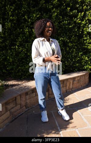 Afrikanische Frau steht in einem Hof an einer Bank und Hecke und lacht, trägt Hemd und Jeans Stockfoto