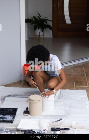Afroamerikanische Frau, die auf einer Abdeckplane an der Haustür kauert und mit einem Werkzeug eine Dose Farbe öffnet, in Jeans-Overalls Stockfoto