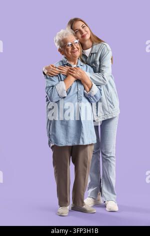 Intergenerationelle Fürsorge und Familienunterstützung, junge Frau umarmt ihre betagte Großmutter mit Liebe und Zuneigung vor einem soliden lila Hintergrund Stockfoto