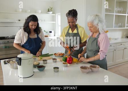 Verschiedene Freundinnen in Schürzen bereiten Mahlzeit an Kücheninsel mit Orangenschäler, Brettern vor. Trio, Kochen, Zusammenarbeit, Zuhause, hell, modern, min Stockfoto