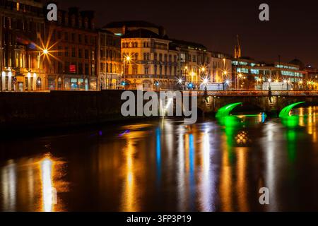 Historische Gebäude am Dubliner Hafen, die nachts neben einer Brücke mit grün beleuchteten Bögen leuchten, mit farbenfrohen Reflexionen auf dem Fluss Liffey und einer Kirche Stockfoto