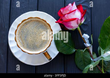 Elegante Tasse frischen Kaffees neben rosa Rose auf dunklem Holztisch, romantisches Stillleben mit Blumendekoration und warmem Getränk. Stockfoto
