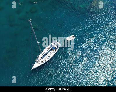 Nahaufnahme aus der Luft mit Drohne eines luxuriösen weißen Segelboots, das in schimmerndem tiefblauem transparentem Meerwasser in Griechenland vor Anker liegt Stockfoto