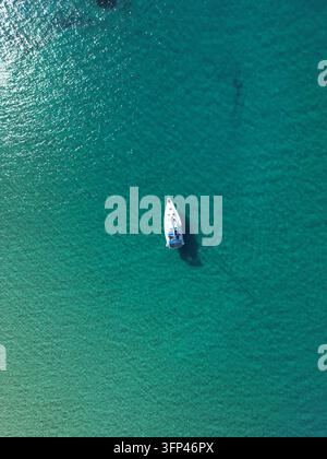 Direkte Overhead-Drohnenaufnahme von einem weißen Segelboot, das in klaren türkisfarbenen Küstengewässern in Griechenland vor Anker liegt. Stockfoto