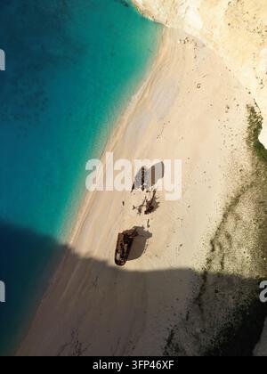 Vertikale Hochwinkel-Drohnenaufnahme von oben, die ein Schiffswrack auf einem weißen Sandstrand zeigt, umgeben von lebhaft blauem Wasser in Griechenland. Stockfoto