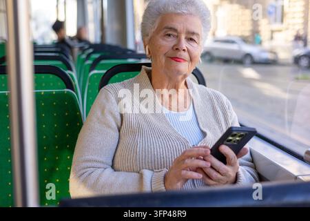 Ältere Frau genießt Audiokontente durch kabellose Ohrhörer während der Stadtfahrt. Seniorenleben, mobile Technologie, öffentliche Verkehrsmittel und aktiv Stockfoto