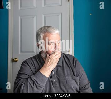 Ein älterer Mann sitzt in einem Raum mit blauen Wänden. Er stützt sein Kinn auf die Hand und schaut zur Seite, versunken in Gedanken. Der Raum hat eine geschlossene Tür in d Stockfoto