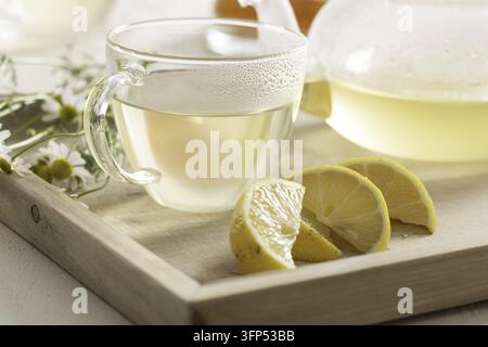 Heißer Kamillentee in einer Glas-Teetasse, die auf einem Holzboden mit Kamillenblüten und Zitrone steht Stockfoto