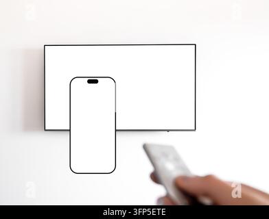 TV-Mockup mit leerem Bildschirm-Handyvorlage, Fernbedienung auf wandmontierten Fernseher gerichtet. Smartphone- und Flachbildschirm-Mockups in der Nähe einer verschwommenen Hand Stockfoto