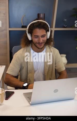 Mann mit weißen Kopfhörern, der auf einen silbernen Laptop schaut, während er am Heimtisch mit einem Smartphone tippt Stockfoto