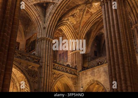 Warmes Licht auf aufragenden gotischen Rippengewölben, gebündelten Steinsäulen und geschnitzten Steinbalkonen in der Kathedrale von Barcelona, mit Gemälden an den Wänden Stockfoto