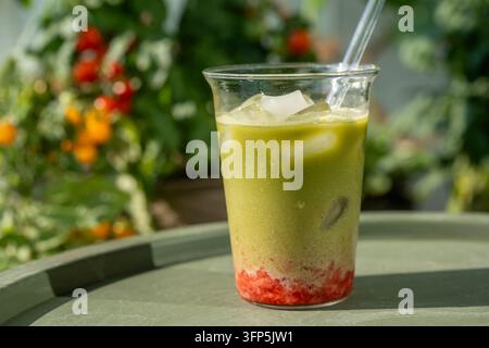 Erfrischendes Eistee- Erdbeer-Matcha-Latte mit bunten Schichten aus frischer Erdbeere, Milch und grünem Matcha, serviert im Freien in einem üppigen Sommergarten Stockfoto