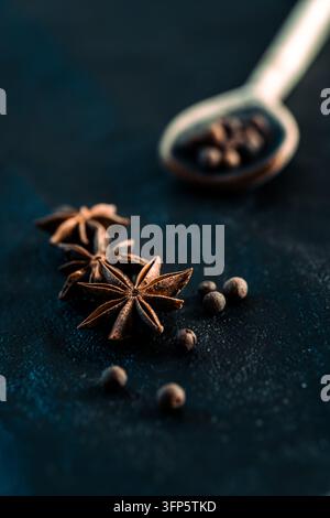 Vertikale Nahaufnahme von getrockneten Sternanisgewürzen und Pfefferkörnern, die auf einer dunklen rustikalen Oberfläche angeordnet sind, mit einem Löffel im Hintergrund. Stockfoto