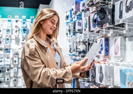 Frau in einem Trenchcoat lächelt, während sie Kopfhörerverpackungen in einem modernen Elektronikgeschäft inspiziert, Optionen vergleicht und den Einkauf von Tech-Zubehör genießt Stockfoto